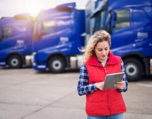 A Woman Checks A Route On A Tablet While Standing In Front Of Trucks Min