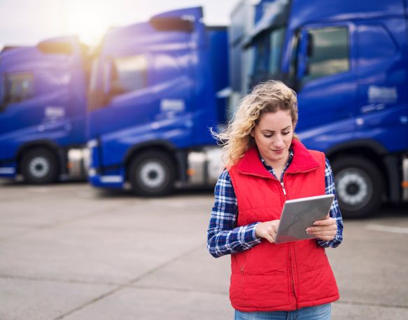 A Woman Checks A Route On A Tablet While Standing In Front Of Trucks Min