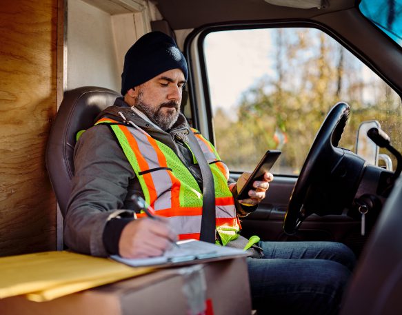 A truck driver taking notes and using mobile phone while sitting in the driver’s seat of his truck