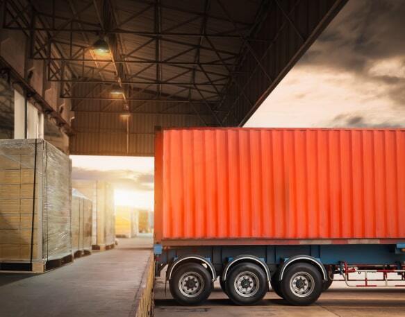 The back of a semi-truck is parked against a shipping warehouse, with pallets positioned next to it.