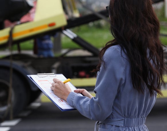 A woman holds a clipboard and takes notes as she watches a trailer being loaded onto a tow truck.