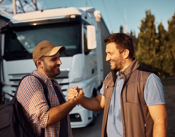 Happy truck driver and his coworker greeting each other while standing in a parking lot.