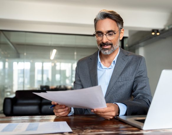 An older male shipping professional works at his desk while looking at some paperwork and smiling.