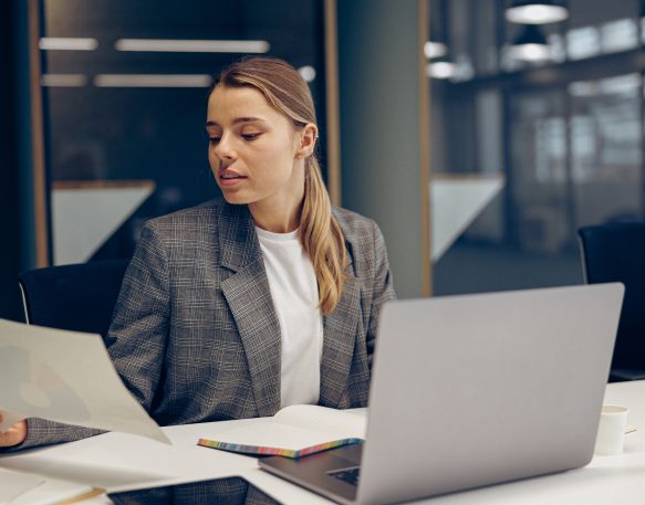 Focused business woman working as a shipper looks at documents and uses a laptop while sitting at a desk in an office.