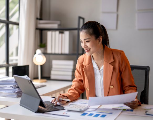 Young female shipper working at desk while looking at papers and scrolling on tablet.
