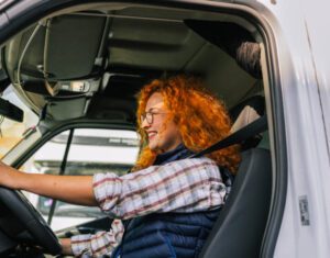A woman with red hair confidently drives a truck, showcasing her role in logistics and transportation
