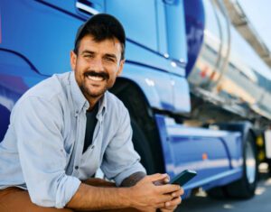 A young male truck driver poses confidently in front of a large, blue truck while holding a cellphone in his hands