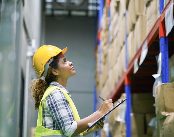 Woman in hard hat and safety vest checking inventory with clipboard in warehouse.