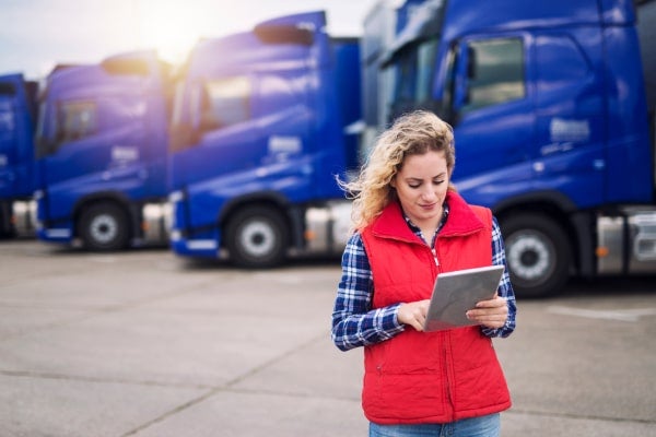 A woman checks a route on a tablet while standing in front of trucks.